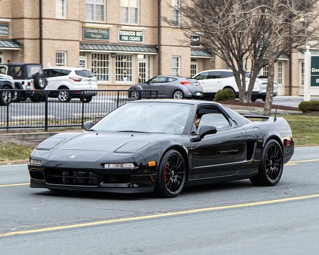 Black Acura NSX (NA1) sports car driving on a street with buildings in background.