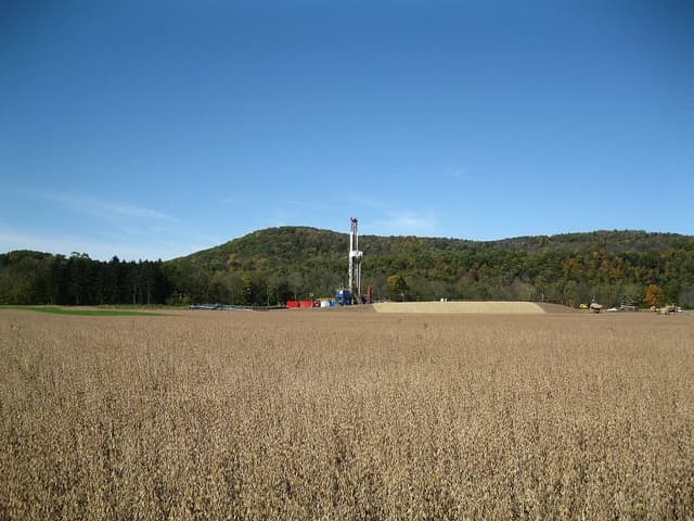 Natural gas well rig operating in a field with a forested hillside in the background.
