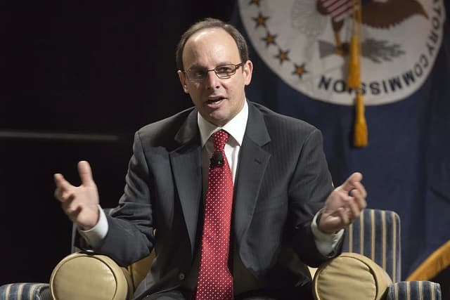 Man in glasses and suit speaking with hands raised at a panel discussion.