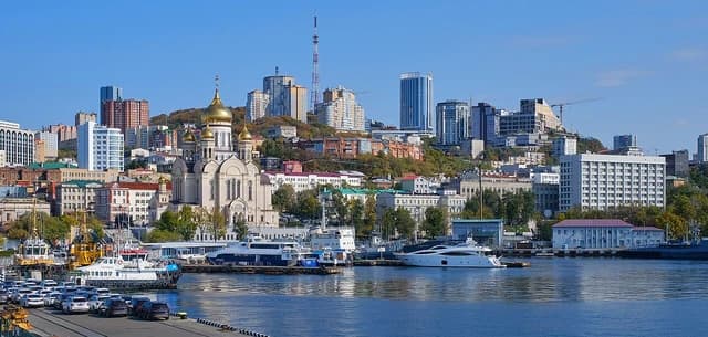 Cityscape of Vladivostok with buildings on hills, a golden-domed church, and boats in the harbor.