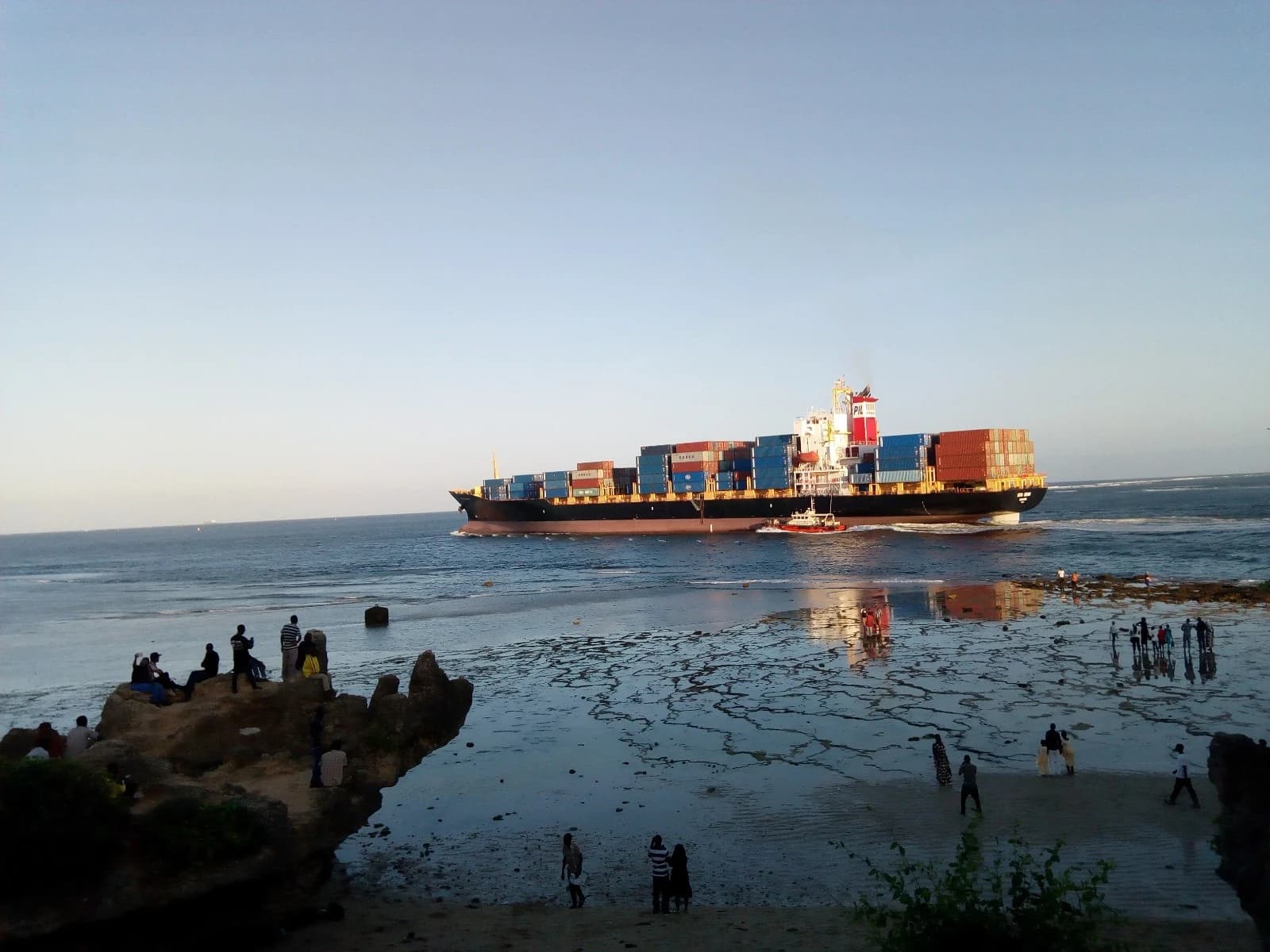 Container ship exits Mombasa port in Kenya, sailing past people on a beach.
