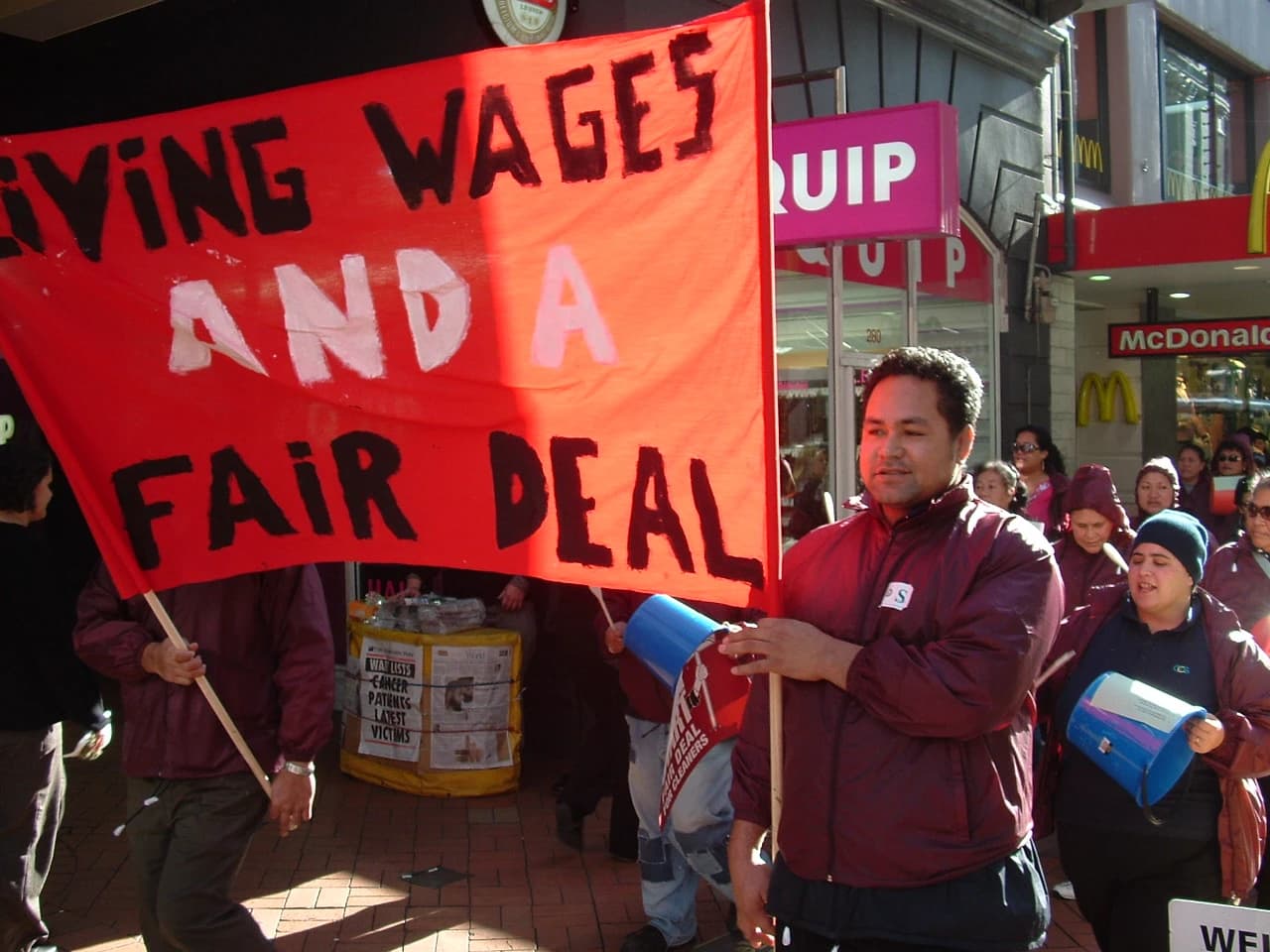Protesters holding a large red banner with 'Living Wages and a Fair Deal' written on it.
