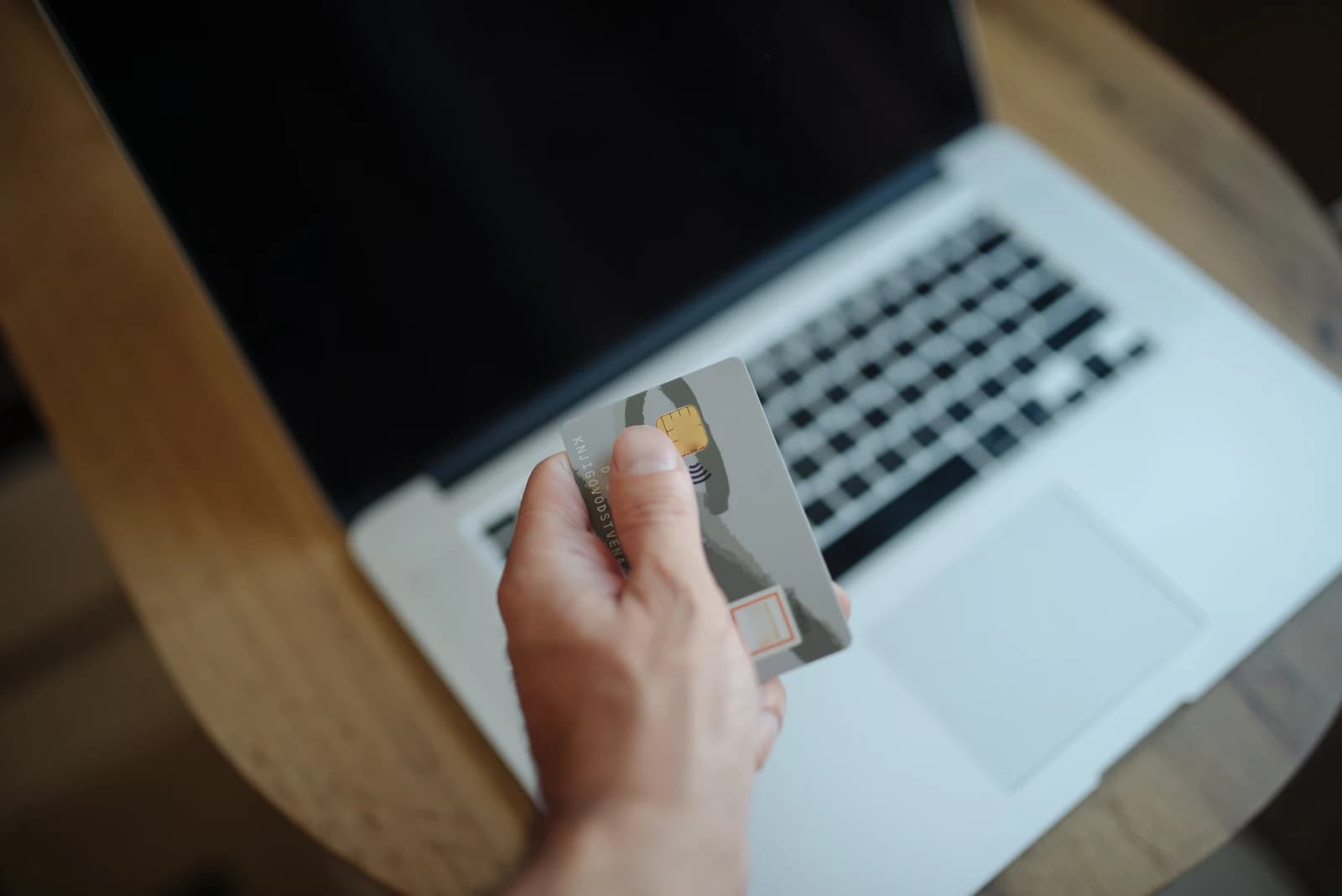 Person holds a credit card while using a laptop on a wooden table.