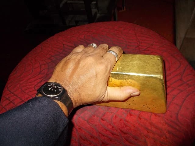 Person's hand with a watch and rings resting on a gold bar.