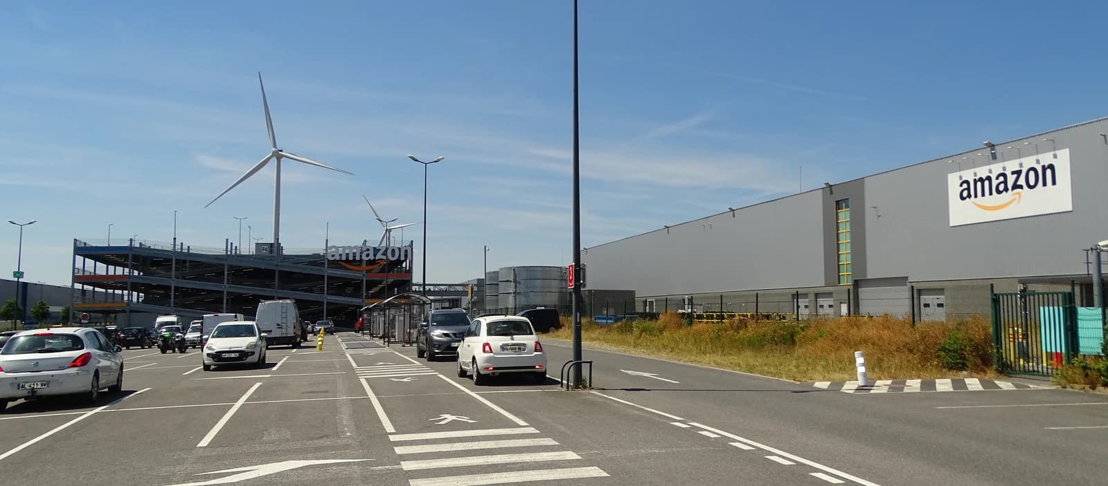 Exterior view of an Amazon logistics center with cars and wind turbines visible.