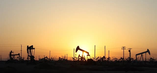 Multiple oil pumpjacks silhouetted against a bright orange and yellow sunset sky.