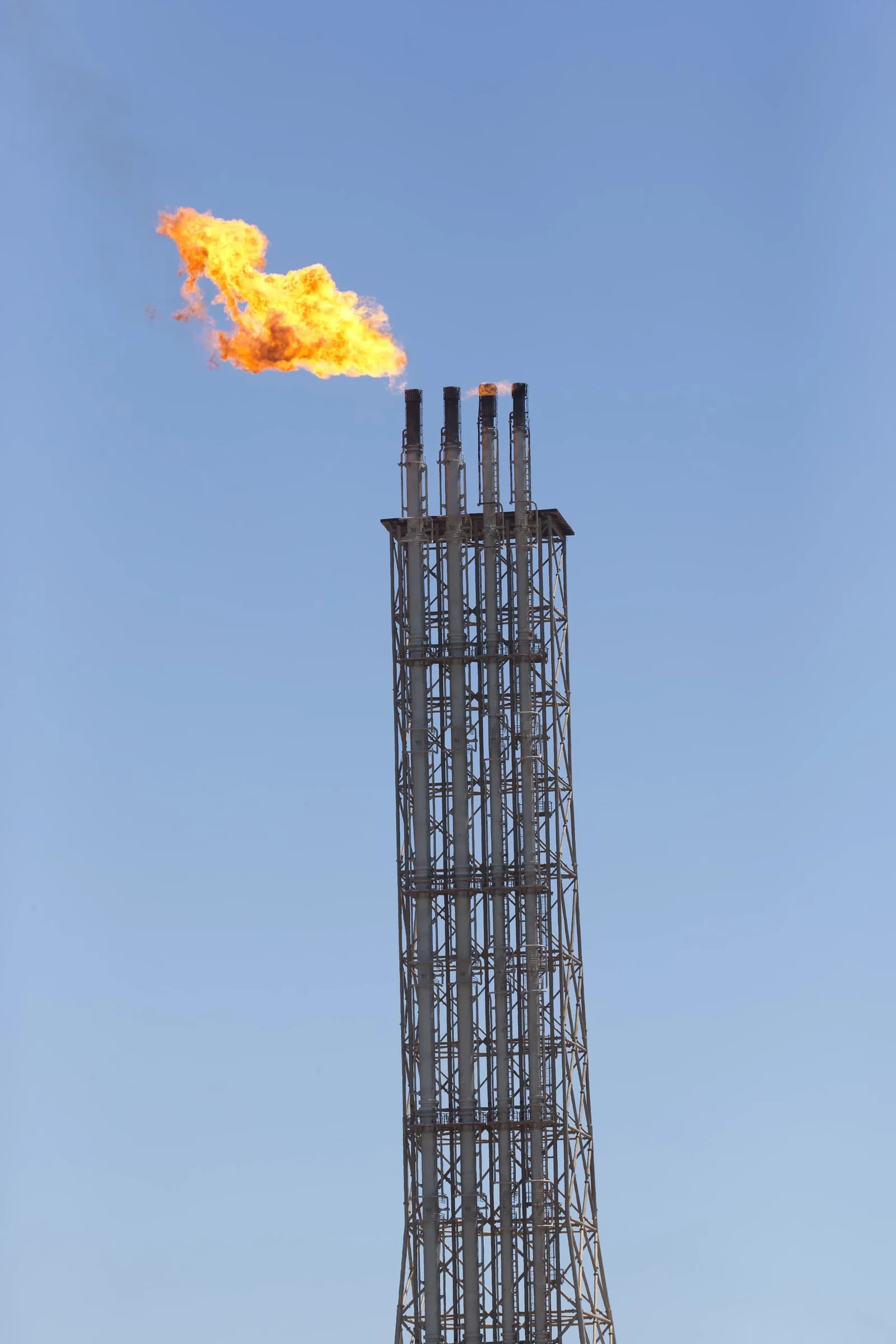 A tall industrial flare stack with a large plume of orange flame burning at its top.