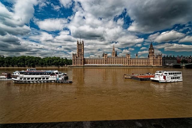 Palace of Westminster, Houses of Parliament, and River Thames with boats under a cloudy sky.
