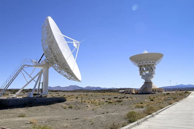 Two large white satellite dishes stand in a desert landscape under a clear blue sky.