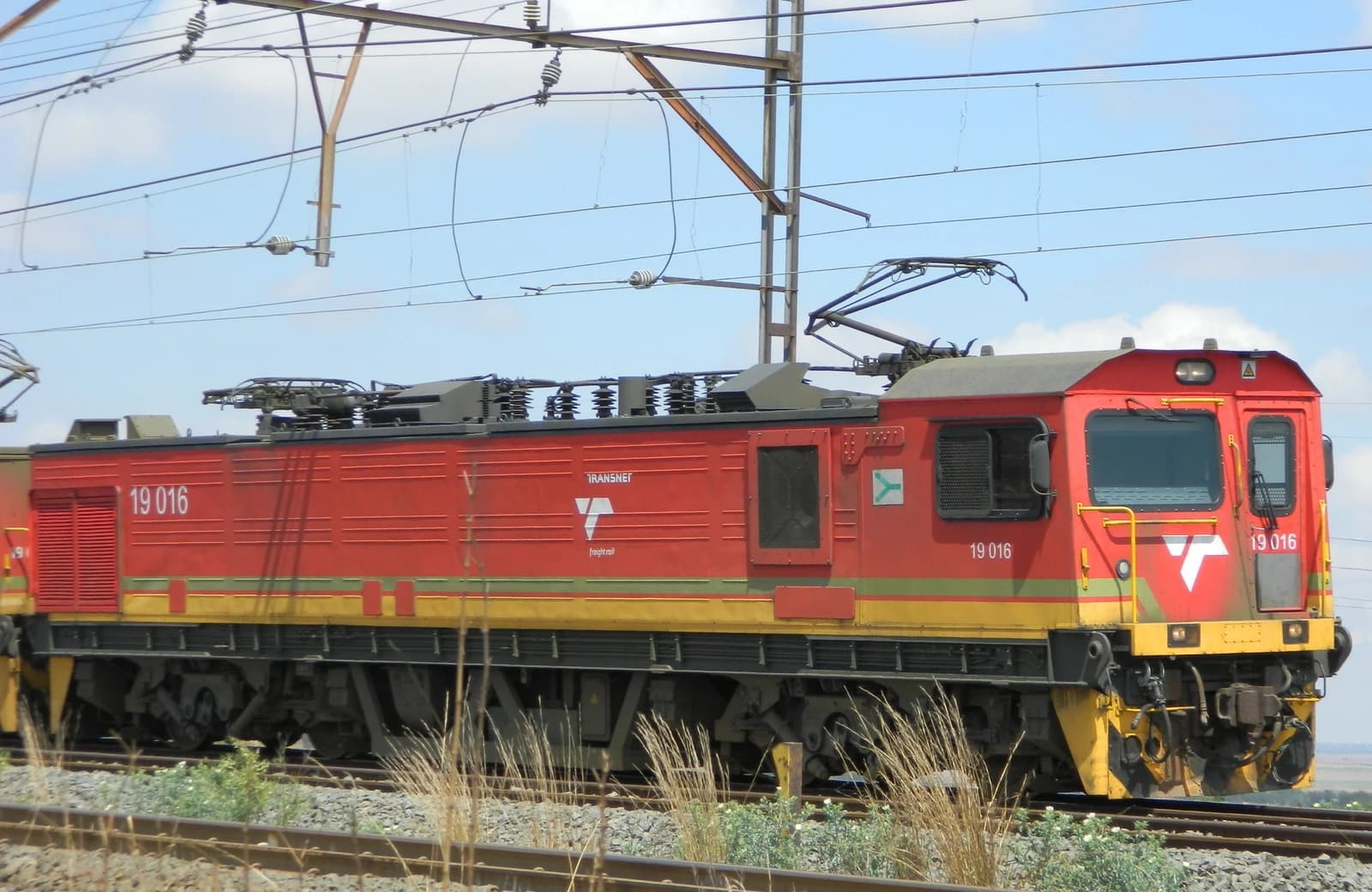 Red and yellow Transnet Class 19-016 locomotive on tracks under overhead lines.