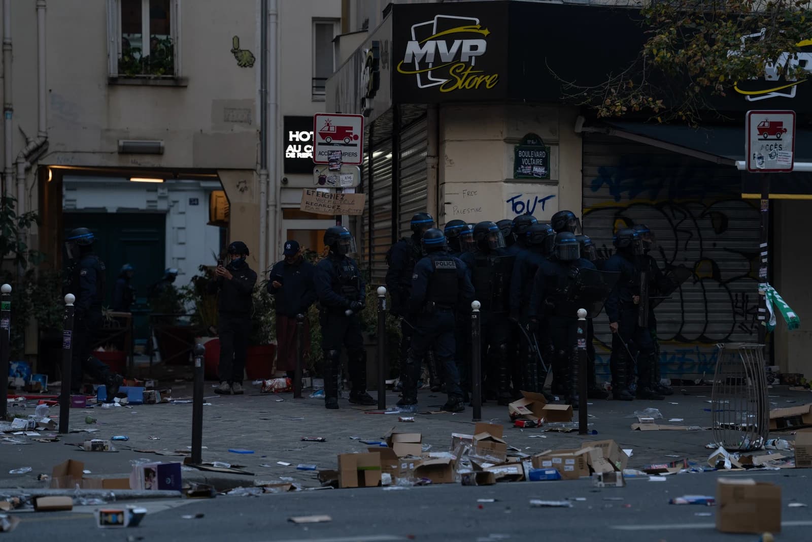 Riot police officers stand on a street littered with debris outside a closed shop.
