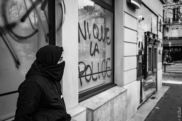 Person in hooded jacket and face covering stands next to a graffiti-covered window.