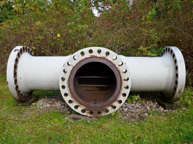 A T-section of a natural gas pipeline sits on the ground with bushes in the background.