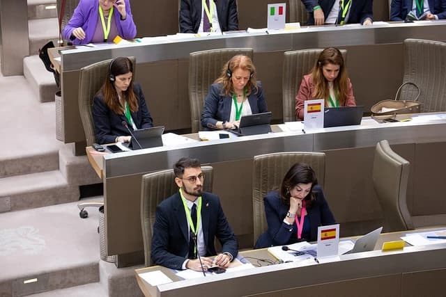 Delegates seated at a conference table with laptops and microphones, representing various countries.