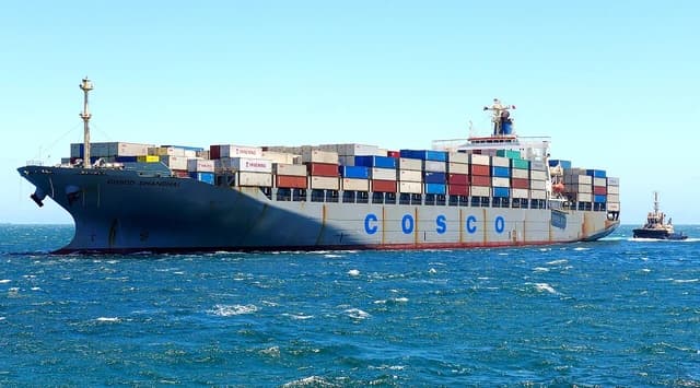 Large container ship 'COSCO Shanghai' with loaded containers in open water, tugboat visible behind.