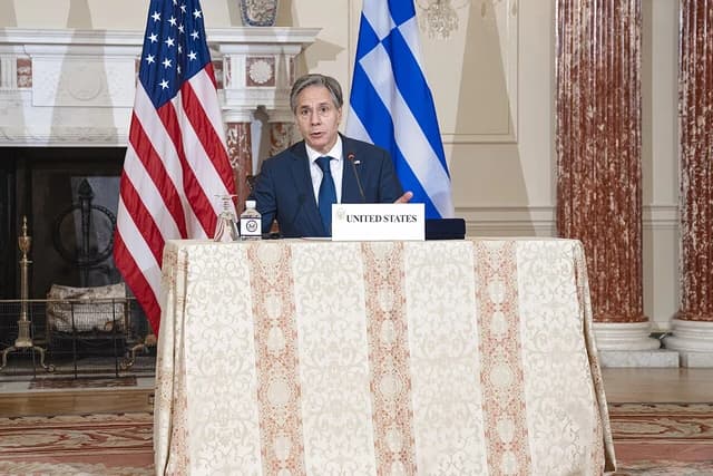 Secretary Blinken speaking at a table with U.S. and Greek flags behind him.