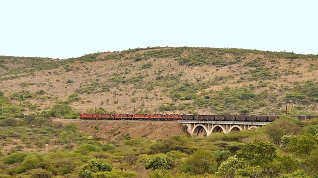 Long freight train with red locomotives crossing a concrete arch bridge in arid hills.
