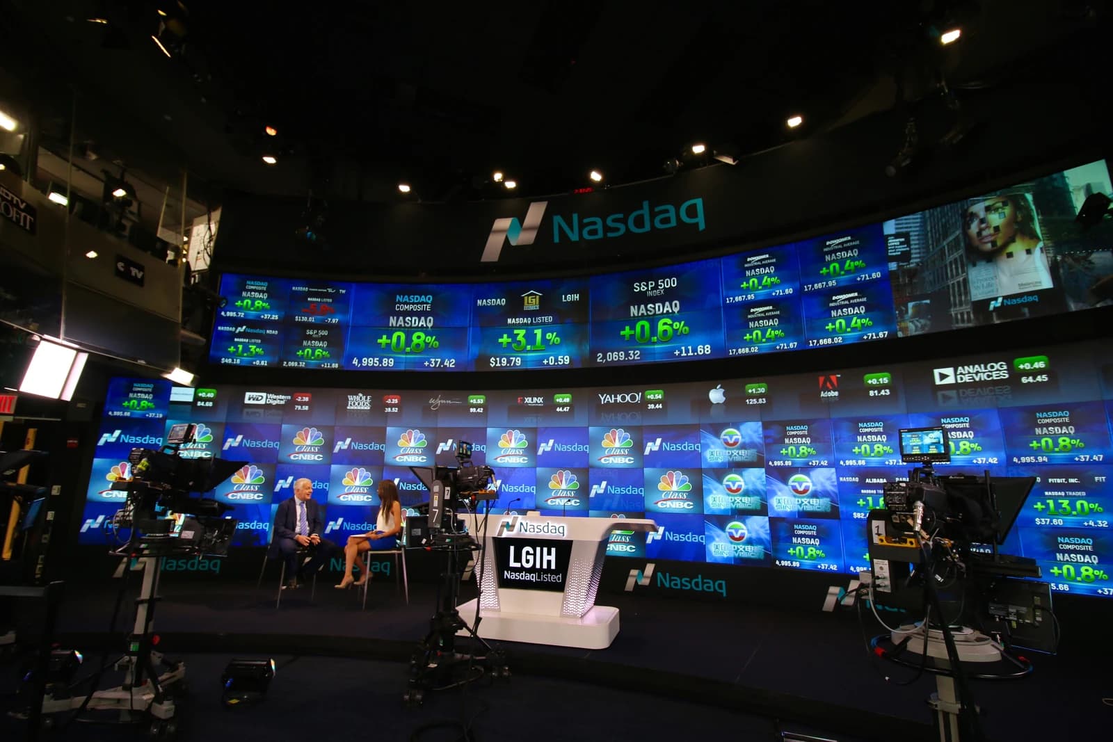 People sit on stage in front of large screens displaying stock market data.
