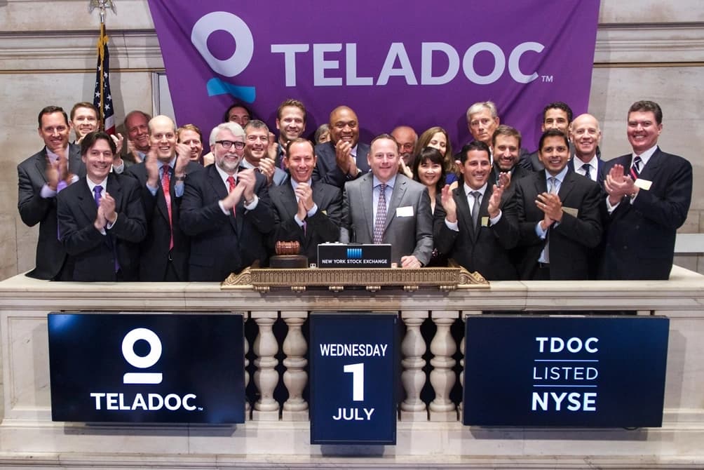 Group of businesspeople, some clapping, at the New York Stock Exchange podium.