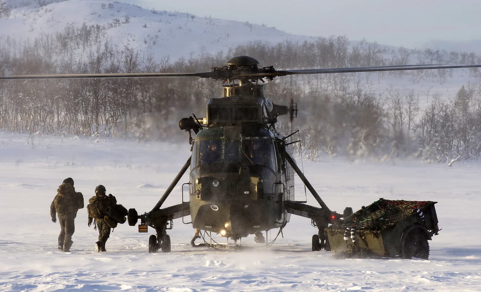 Two military personnel walk toward a Royal Navy Sea King Mk4 helicopter on a snowy field.