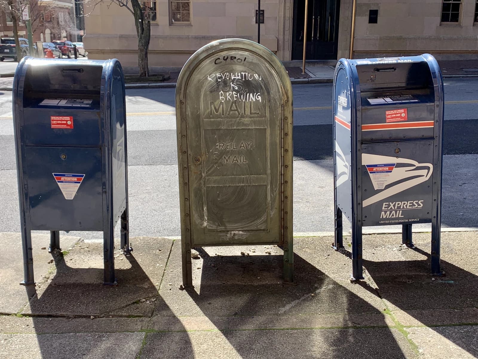 Three different United States Postal Service mailboxes standing on a city sidewalk.
