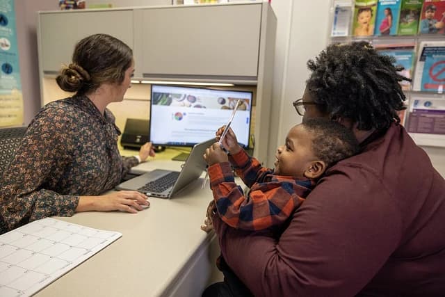 A mother holds a young child while speaking with a WIC program employee at a desk.