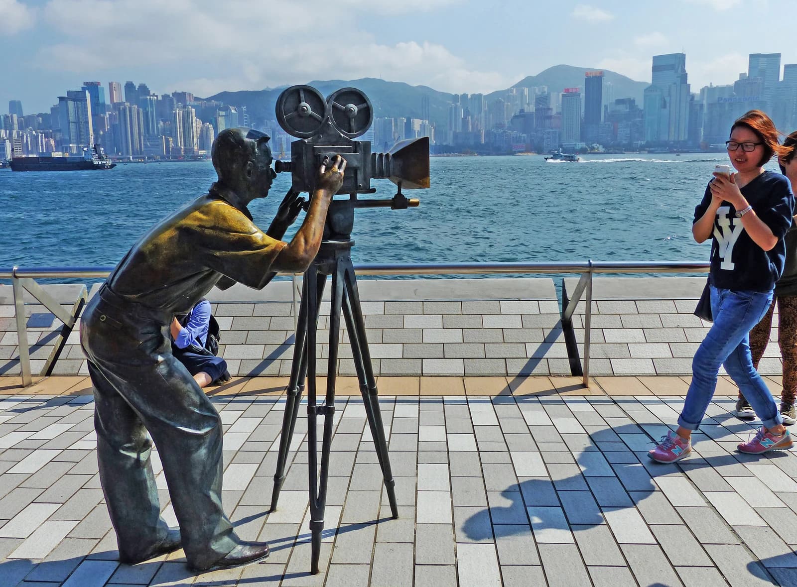 Bronze statue of a cameraman operating an old film camera by a waterfront with a city skyline.