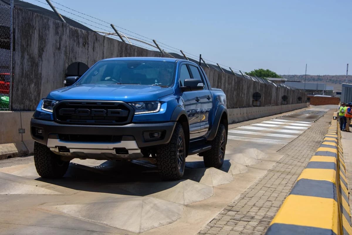 Blue Ford Ranger Raptor pickup truck driving over a concrete obstacle in daylight.
