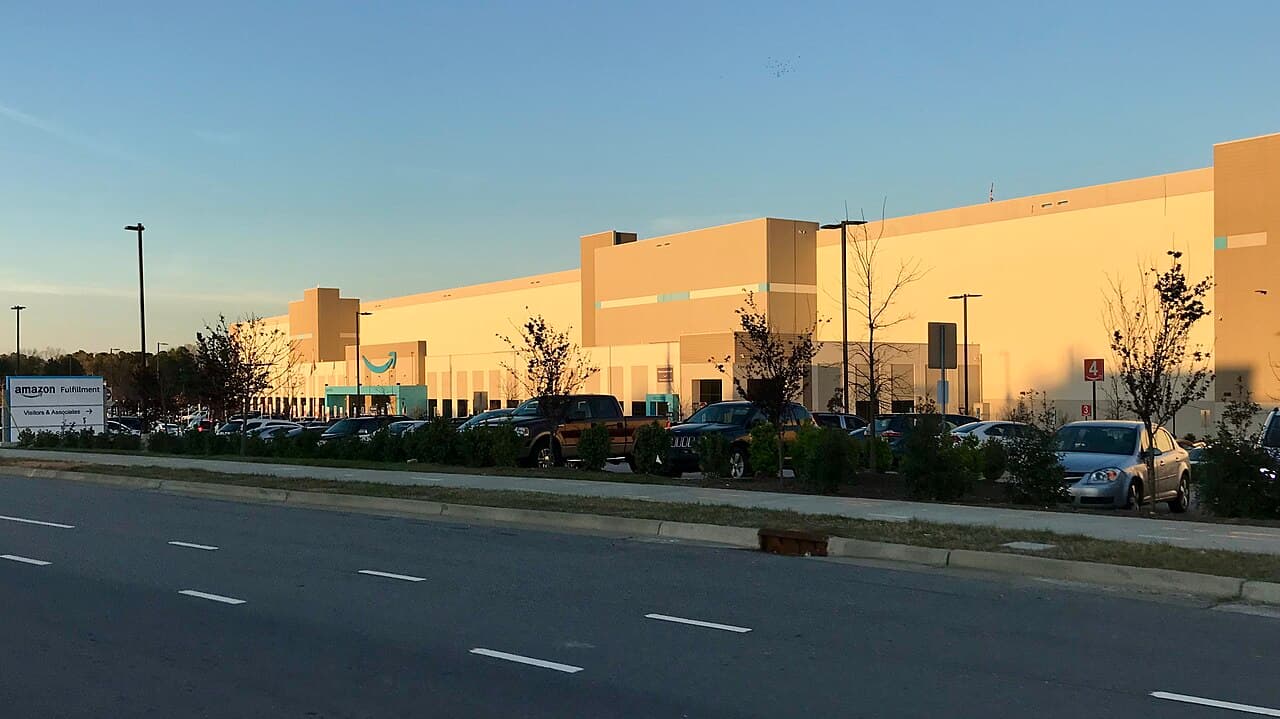 Exterior view of an Amazon warehouse building with a parking lot and road in front.