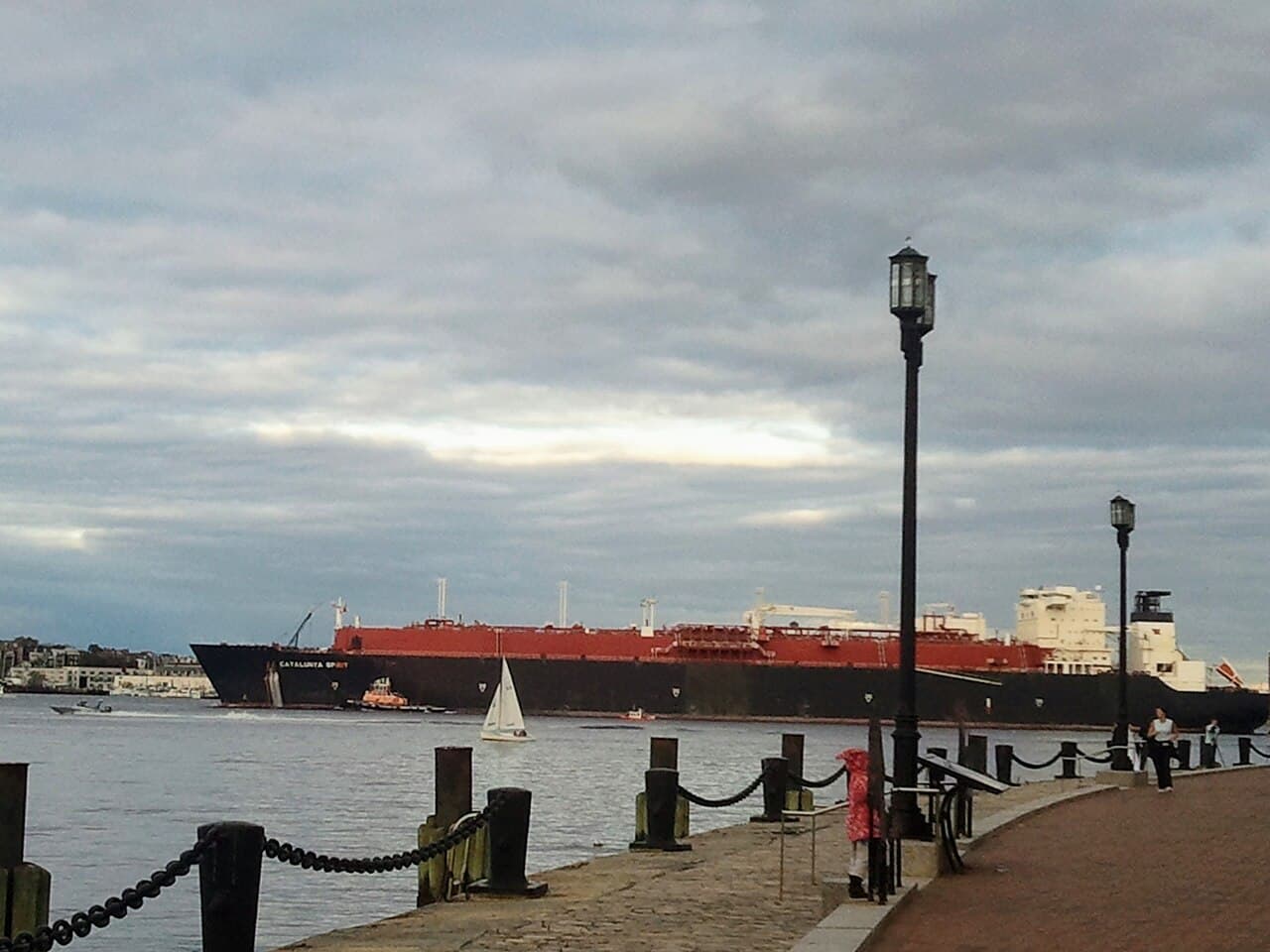 Large red and black LNG tanker 'Catalunya Spirit' sailing in Boston harbor with a sailboat nearby.